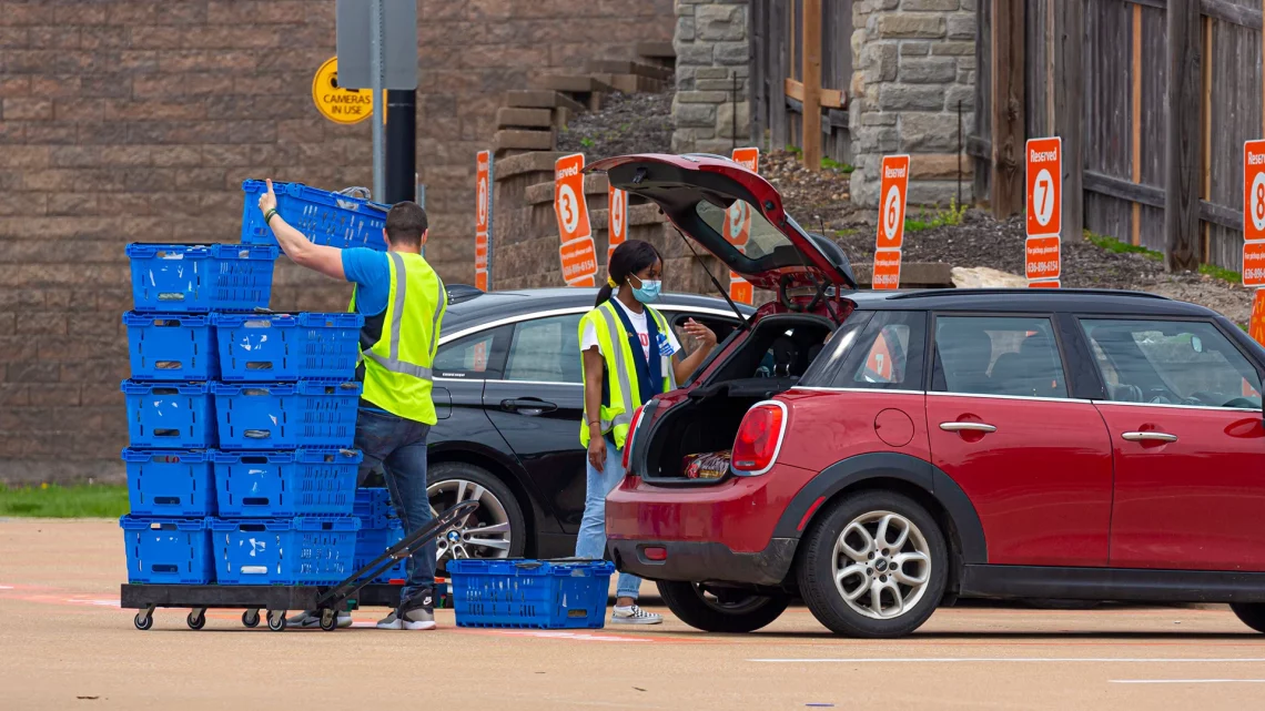 People in parking lot loading cart-picked groceries into car during Covid